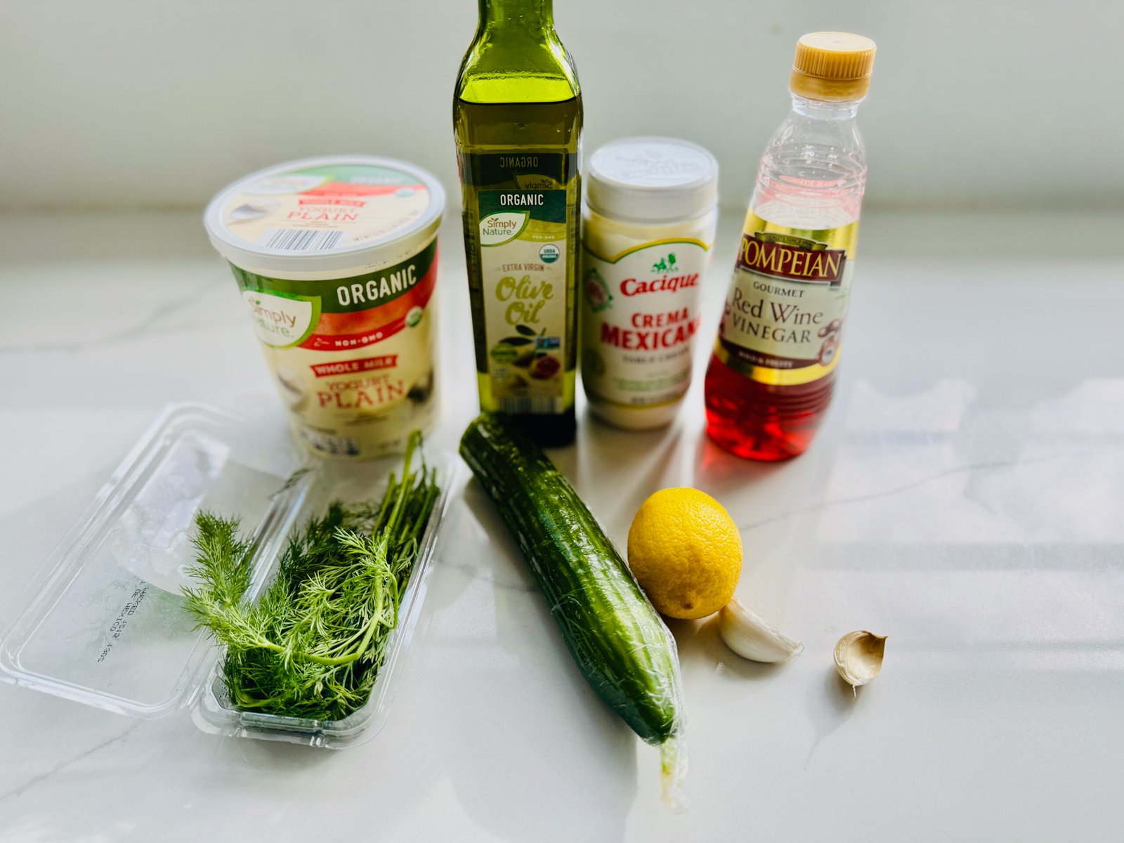 Ingredients for creamy Greek sauce including yogurt, olive oil, crema mexicana, cucumber, lemon, garlic, dill, and red wine vinegar