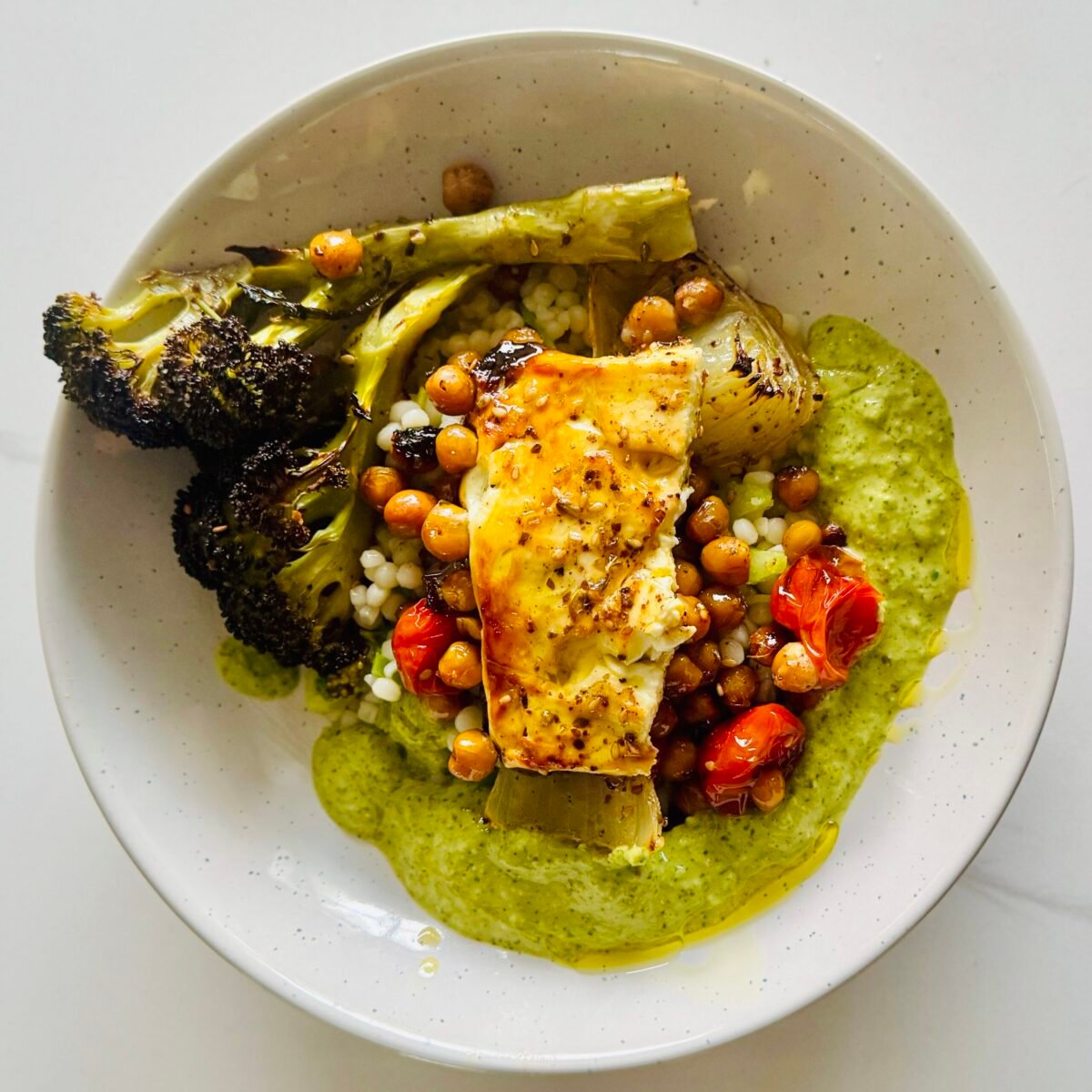 baked feta bowl with chickpeas broccoli tomatoes couscous and green sauce in a shallow serving bowl