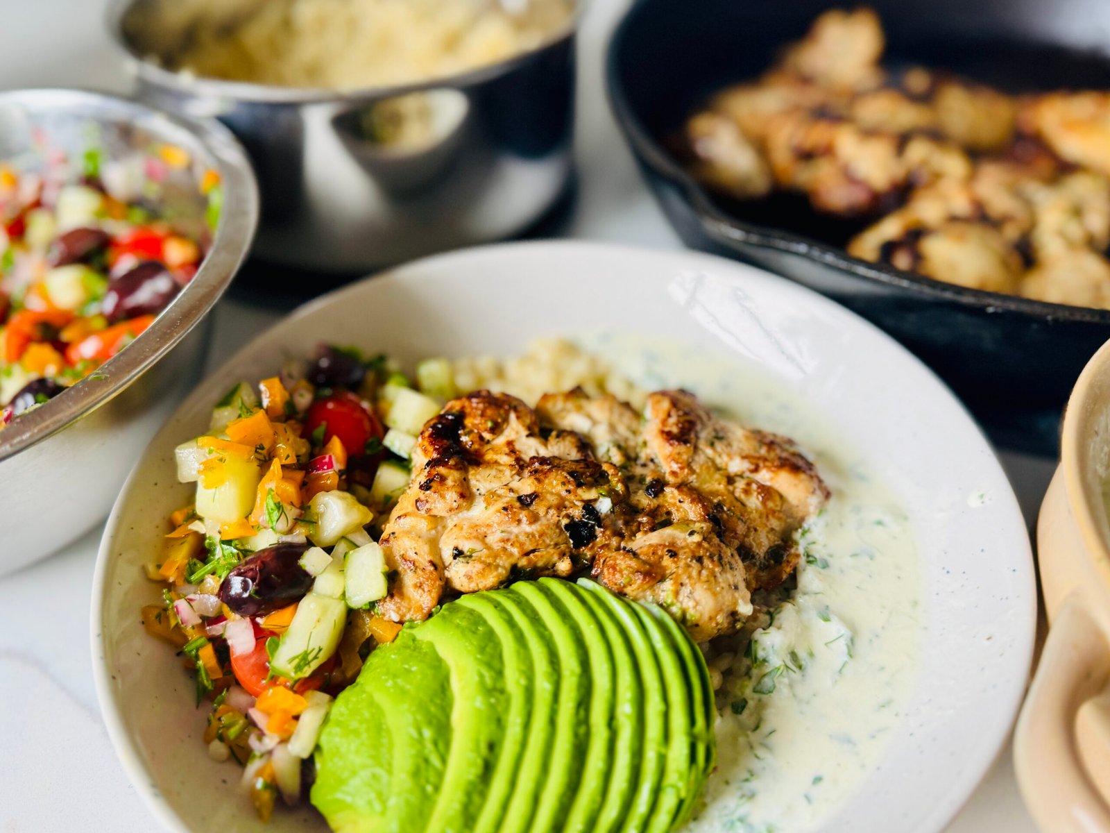 Greek chicken bowl with yogurt marinated chicken, garlic couscous, cucumber, tomatoes, olives, and creamy Greek sauce