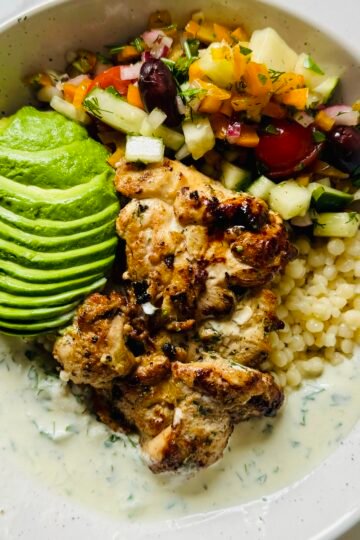 Greek chicken bowl with yogurt marinated chicken, garlic couscous, cucumber, tomatoes, olives, and creamy Greek sauce