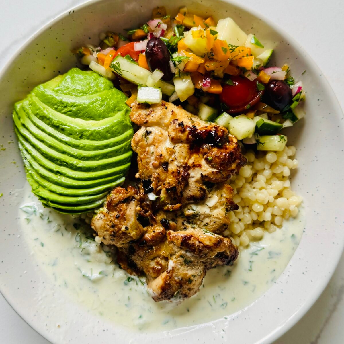 Greek chicken bowl with yogurt marinated chicken, garlic couscous, cucumber, tomatoes, olives, and creamy Greek sauce