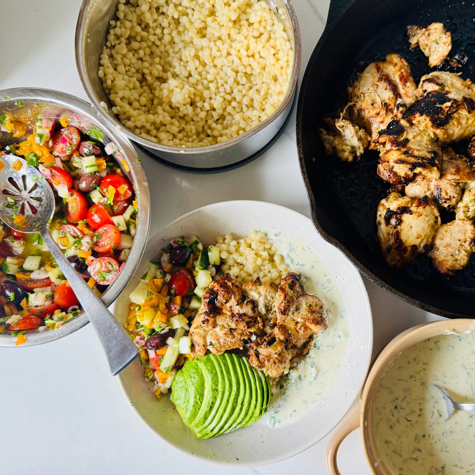 Greek chicken bowl with yogurt marinated chicken, garlic couscous, cucumber, tomatoes, olives, and creamy Greek sauce