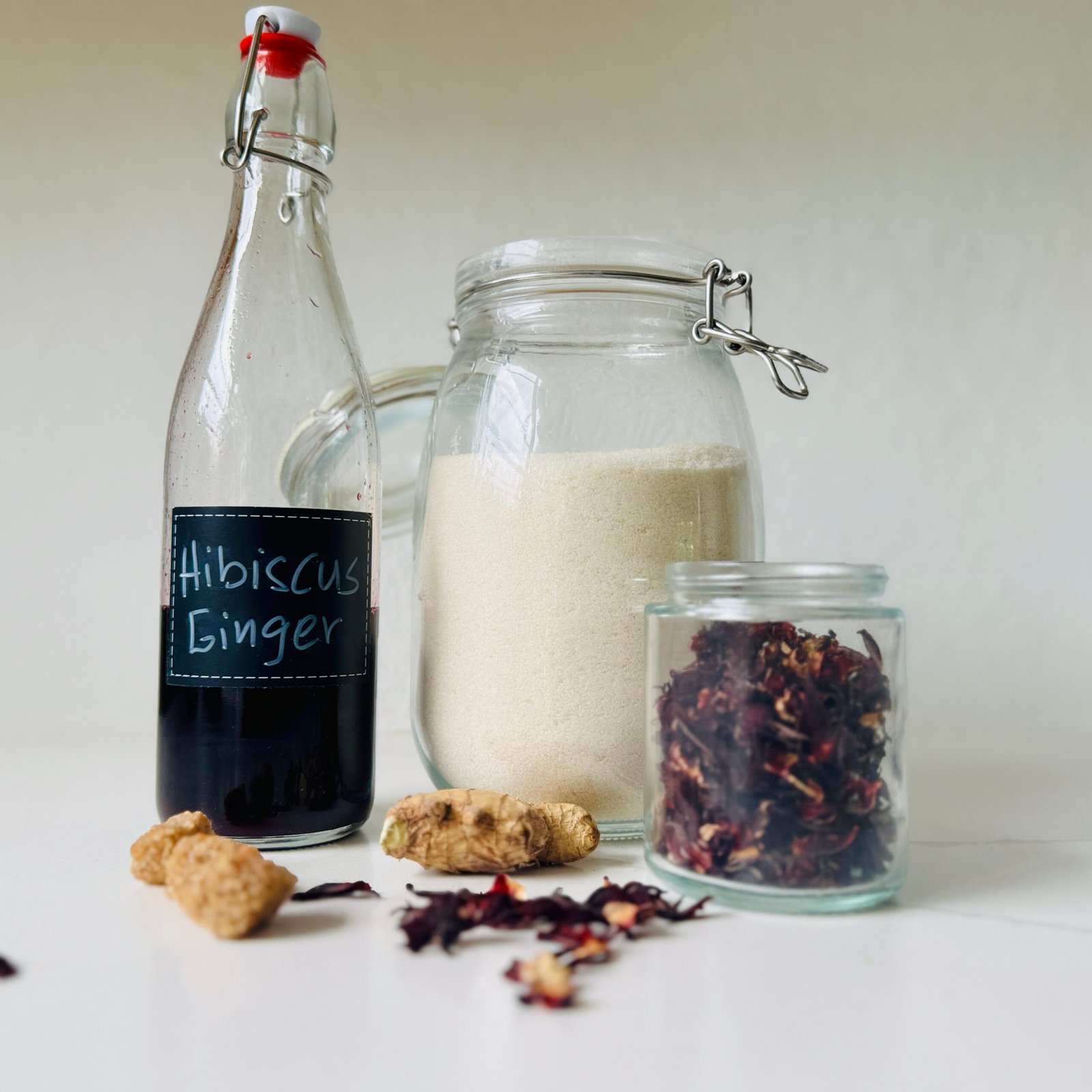 Dark red hibiscus-ginger syrup in a labeled bottle with dried hibiscus, fresh ginger, and sugar.