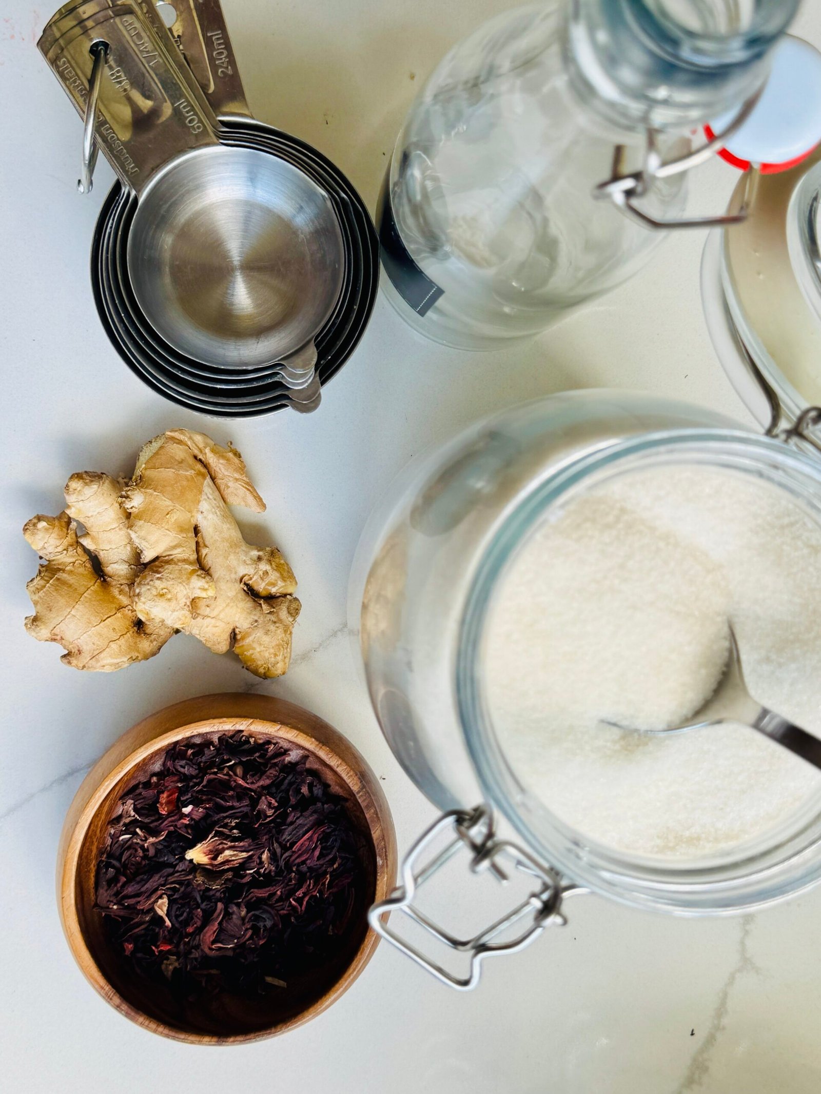Dried hibiscus flowers, fresh ginger, granulated sugar, measuring cups, and a glass bottle for hibiscus-ginger syrup.