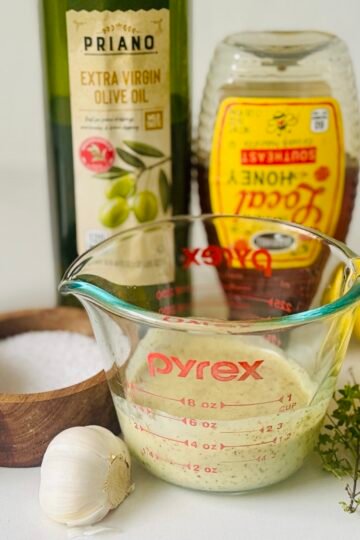 Lemon Dijon marinade in a glass measuring cup beside garlic, lemon, herbs, olive oil, and honey.