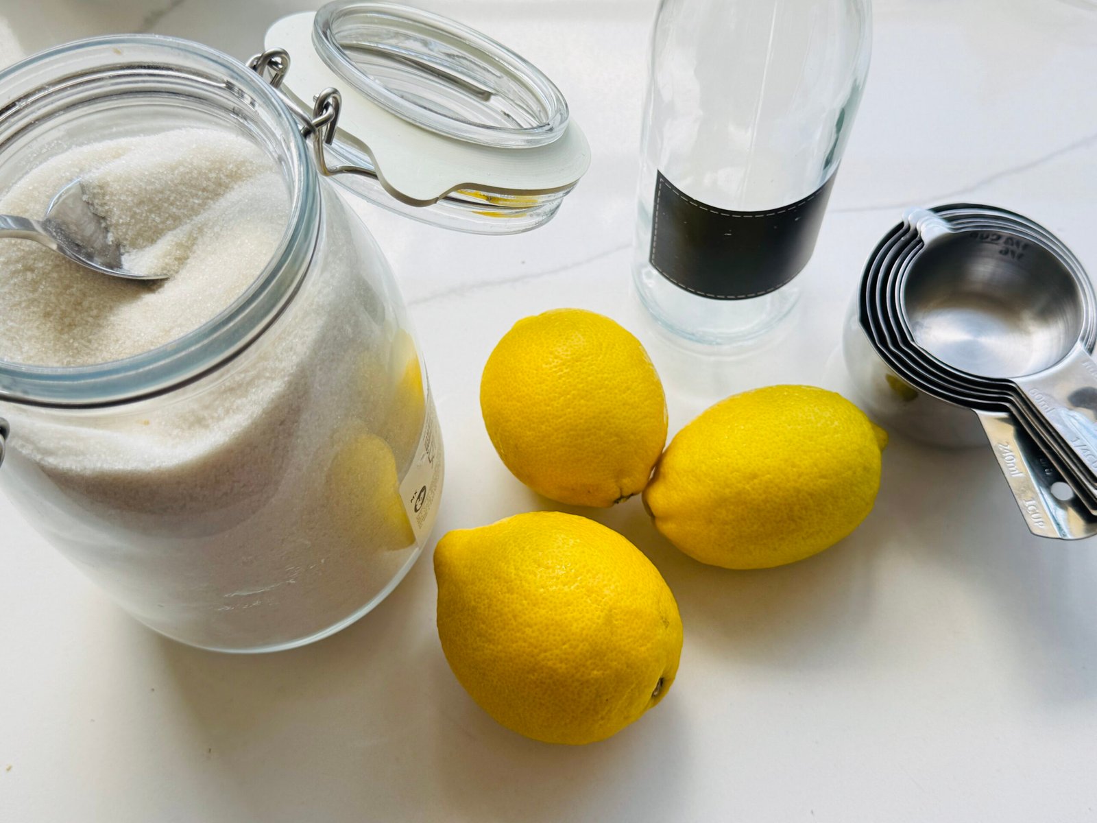 Fresh lemons, granulated sugar, measuring cups, and a glass bottle for lemon oleo-saccharum.