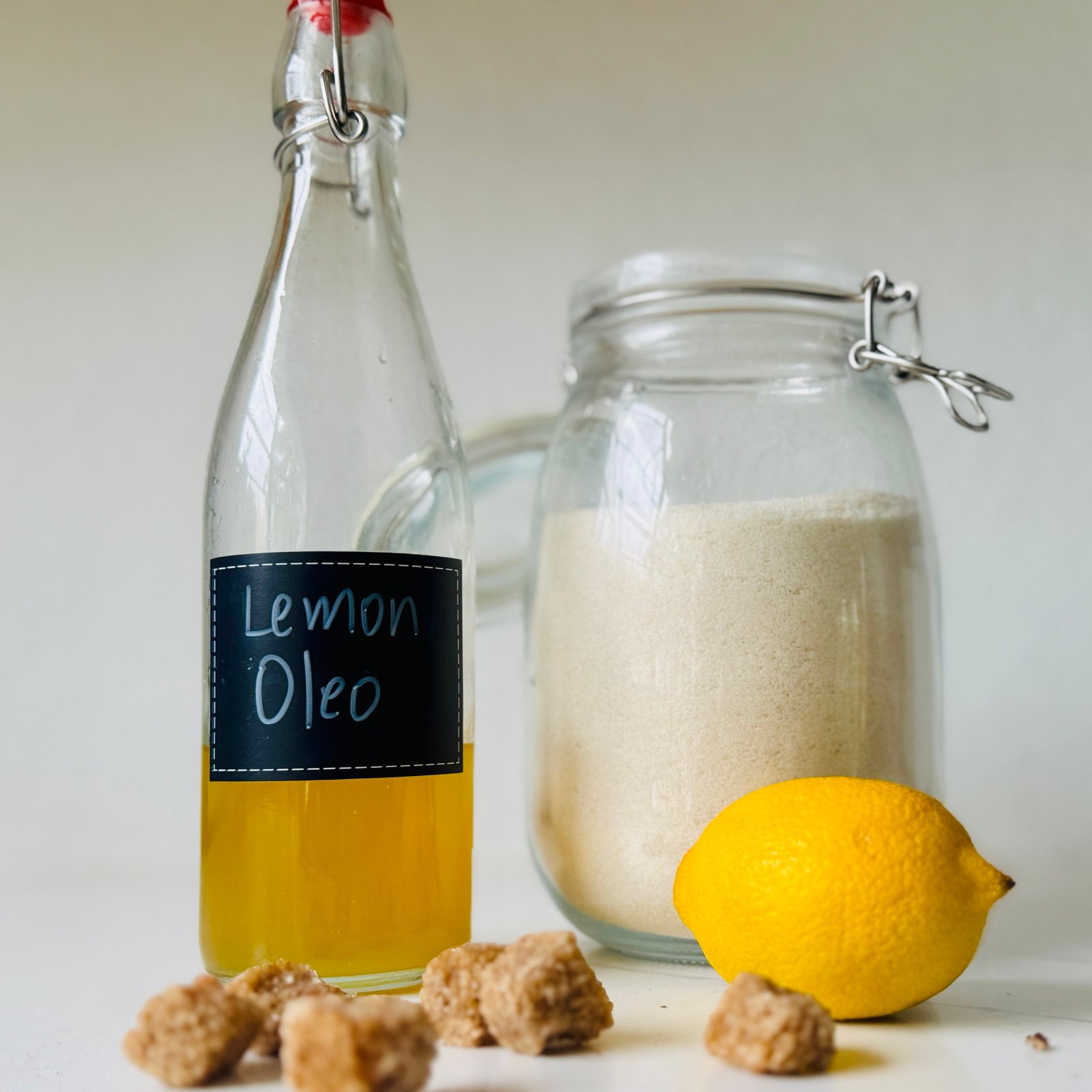 Golden lemon oleo-saccharum syrup in a labeled bottle beside a lemon and granulated sugar.