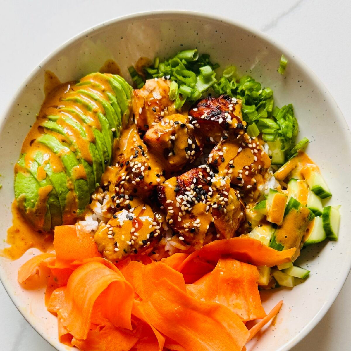 Soy ginger glazed salmon rice bowl with jasmine rice, cucumber, carrot ribbons, avocado, scallions, sesame seeds, and creamy gochujang sauce