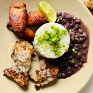 Jerk chicken with creamy coconut sauce served beside coconut rice topped with scallions, black beans, caramelized plantains and a lime wedge.
